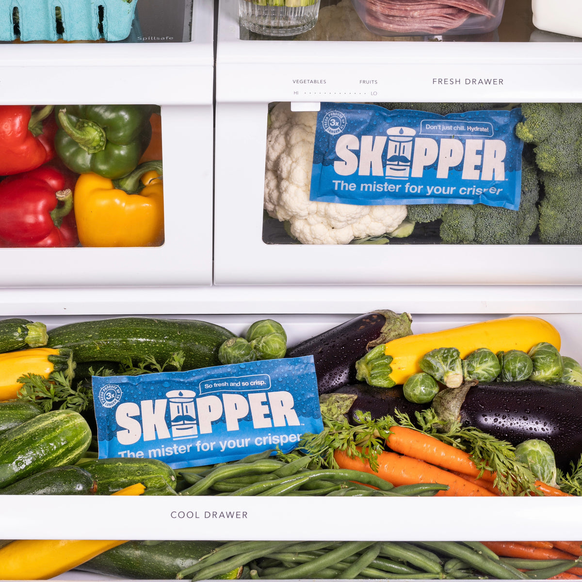 Refrigerator crisper drawers filled with bell peppers, cauliflower, broccoli, zucchini, eggplant, carrots, and Brussels sprouts, with two Skipper packets placed among the produce.