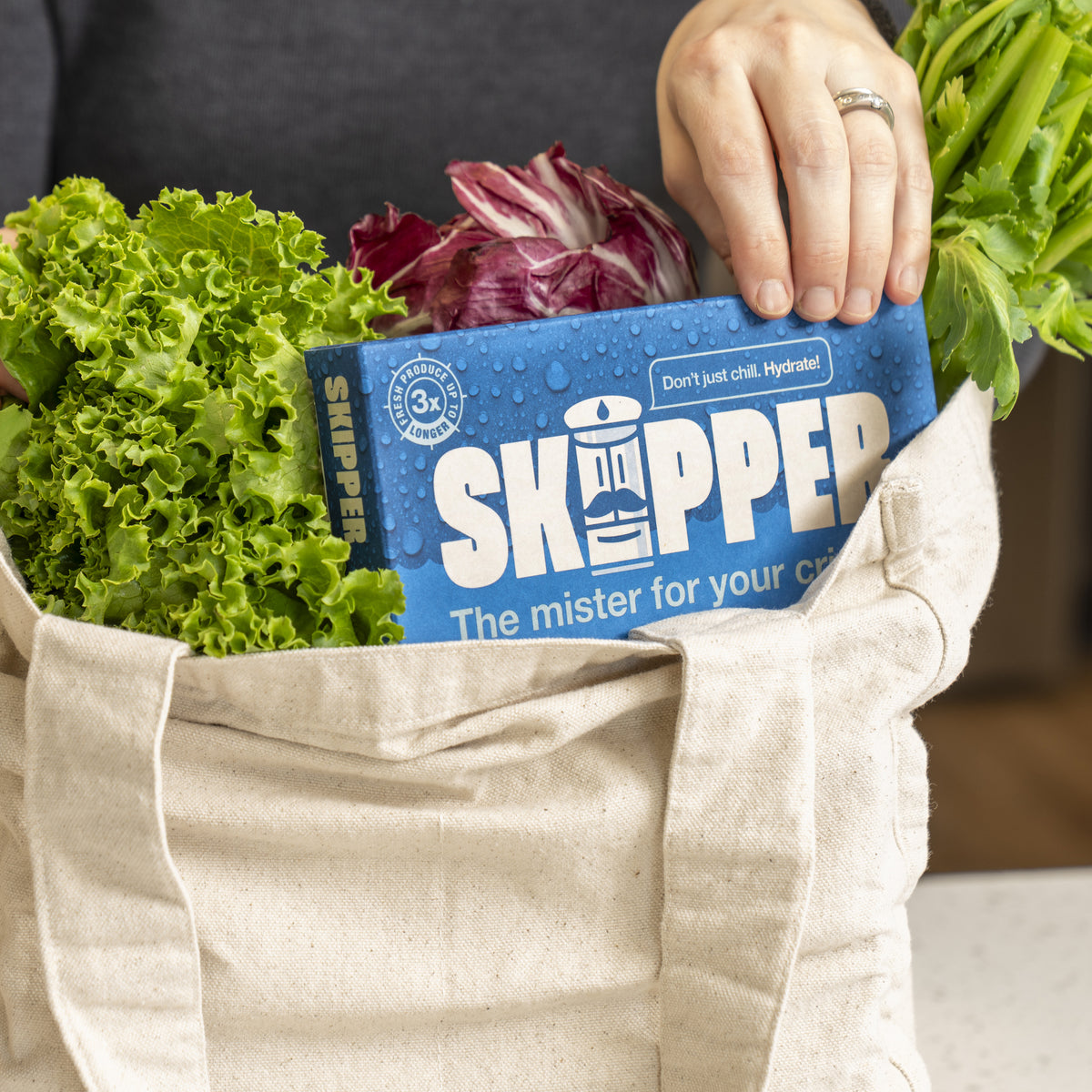 Hand placing a Skipper box into a canvas tote bag filled with fresh lettuce, celery, and radicchio.